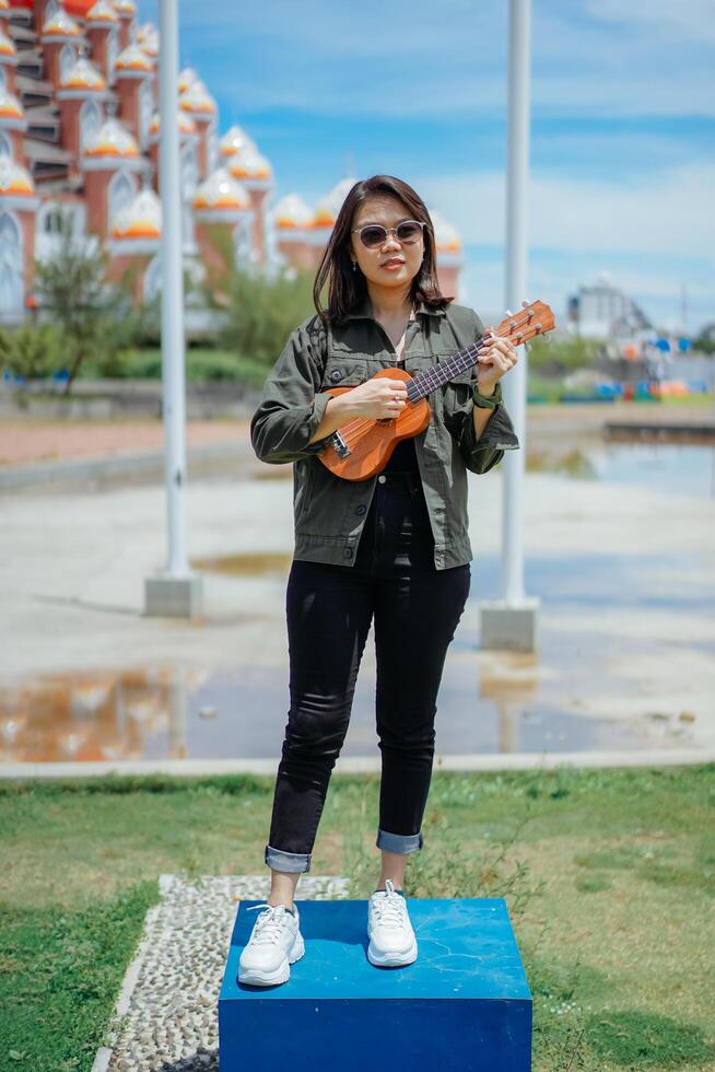 spelar ukulele av ung skön asiatisk kvinna bär jacka och svart jeans Framställ utomhus foto