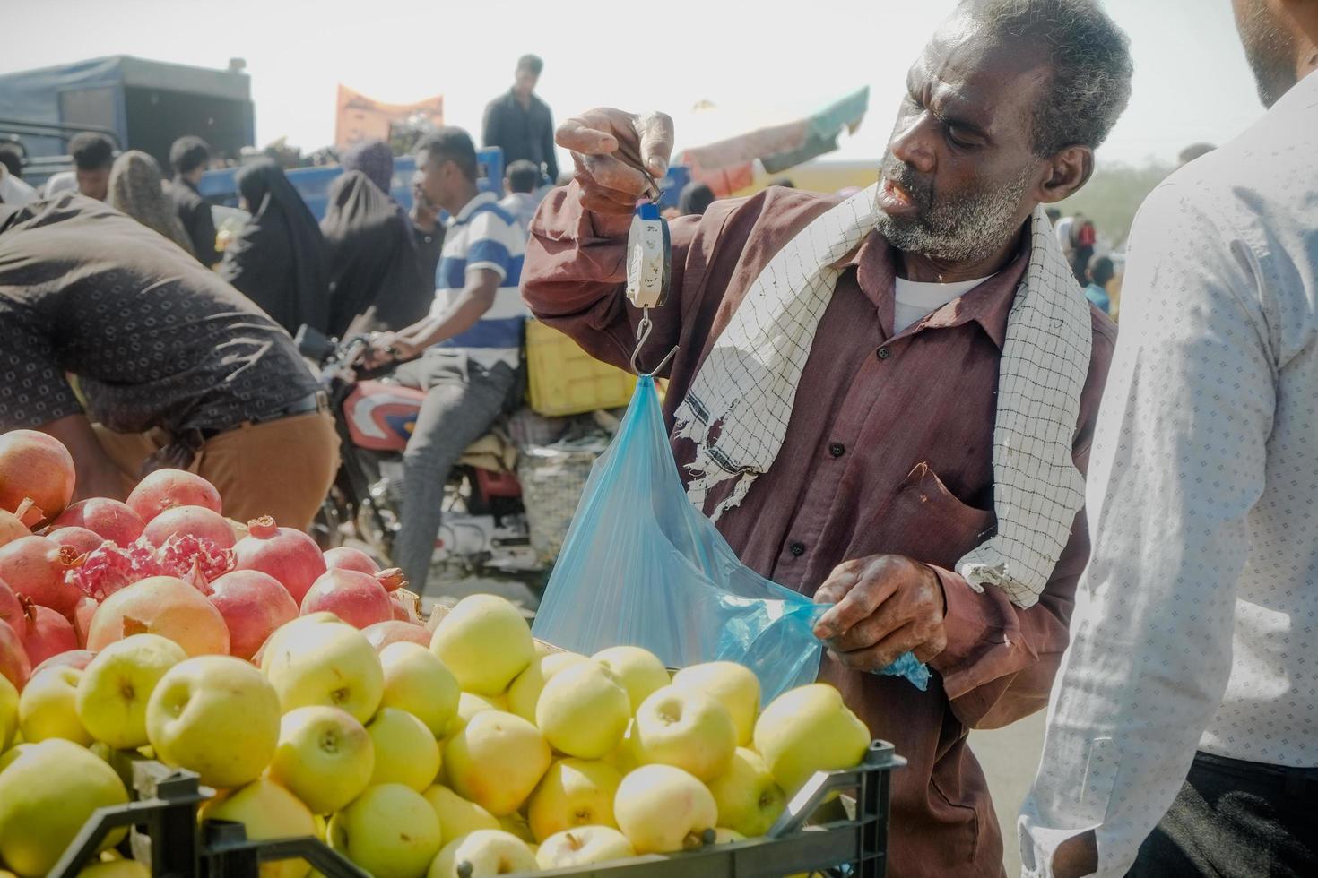Hormozgan -provinsen, Iran, 2016 - lokal iransk gatufruktförsäljare som använder manuell hängande vägningsvåg för att sälja varor på sitt lilla företag på minab torsdag färskmarknad foto