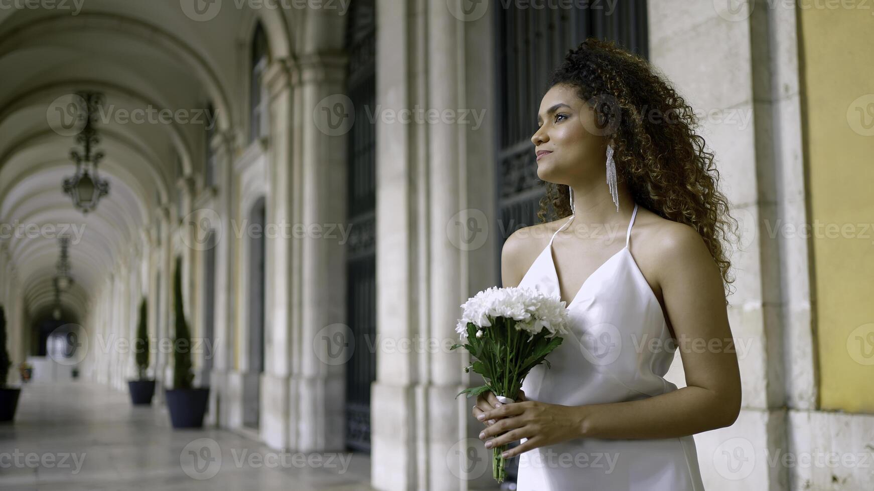 skön svart kvinna i elegant vit klänning. handling. attraktiv kvinna i vit klänning med bukett på gammal byggnad. attraktiv ung kvinna är väntar för pojkvän med blommor foto