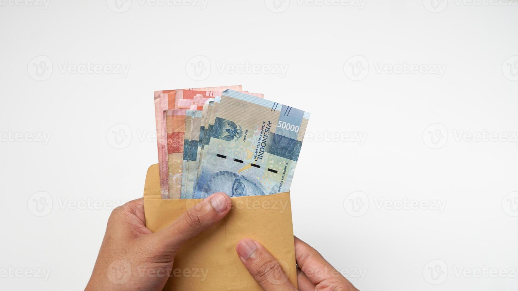 både händer är innehav en brun kuvert och indonesiska rupiah sedlar på en vit bakgrund foto