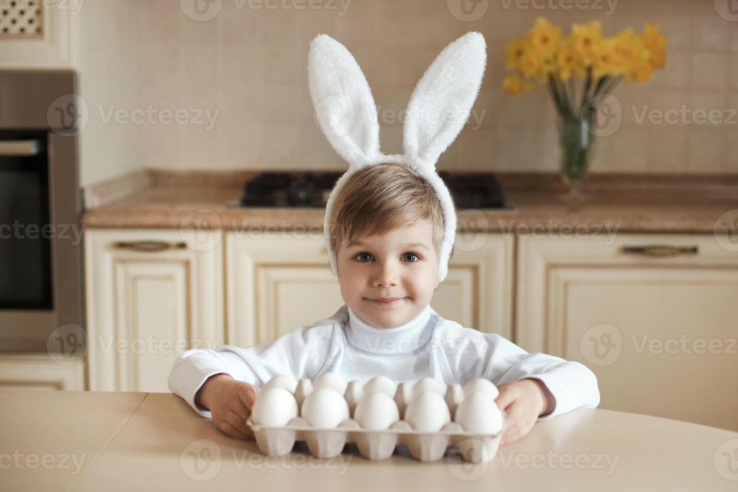 söt caucasian unge bär kanin öron och innehav uppsättning av vit organisk chiken ägg, framställning till påsk. liten pojke Sammanträde på tabell och ser på kamera. foto