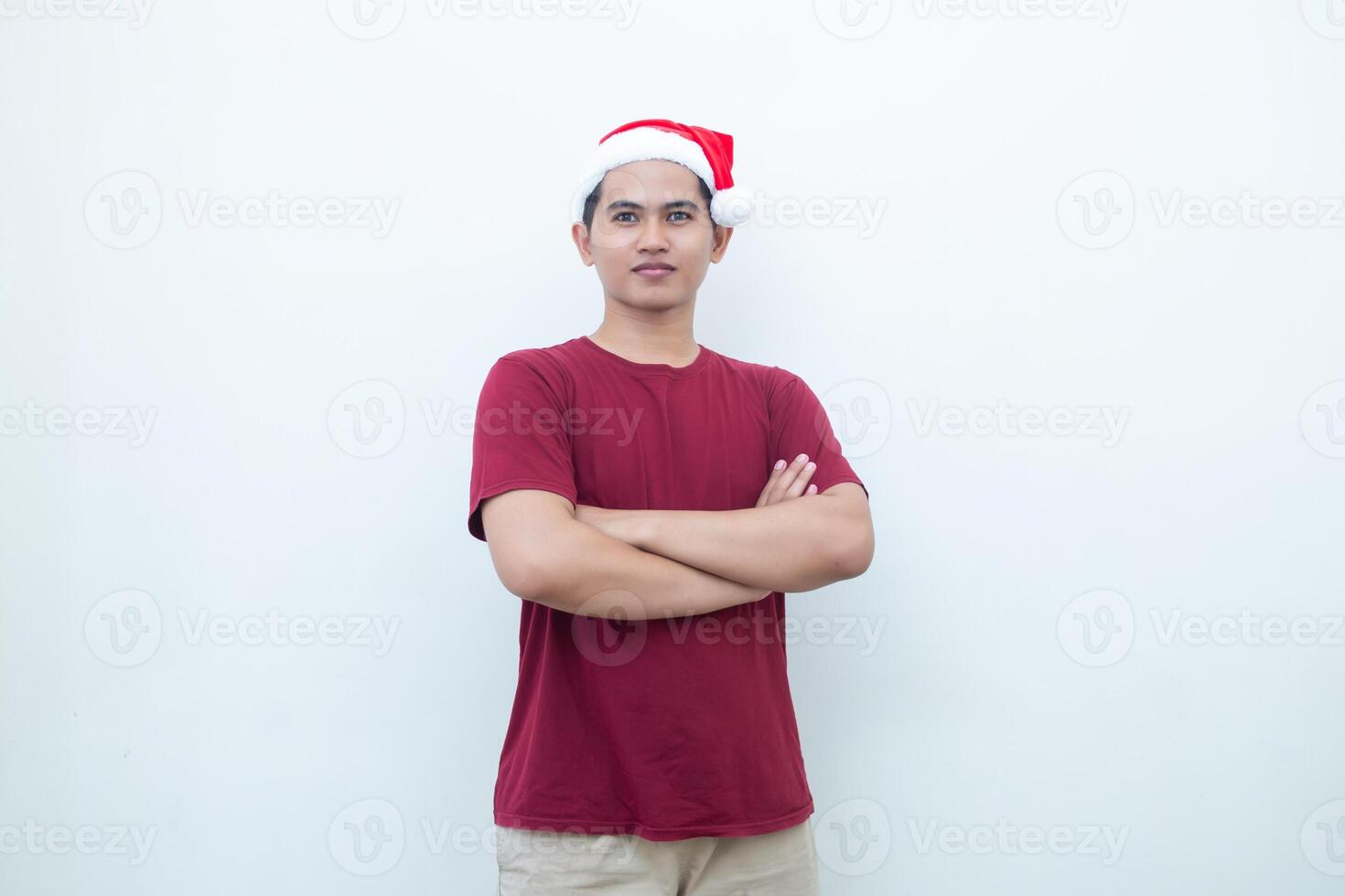 ung asiatisk man bär en santa claus hatt korsning hans vapen medan leende, chock och skrattande med en vit bakgrund för visuell kommunikation foto