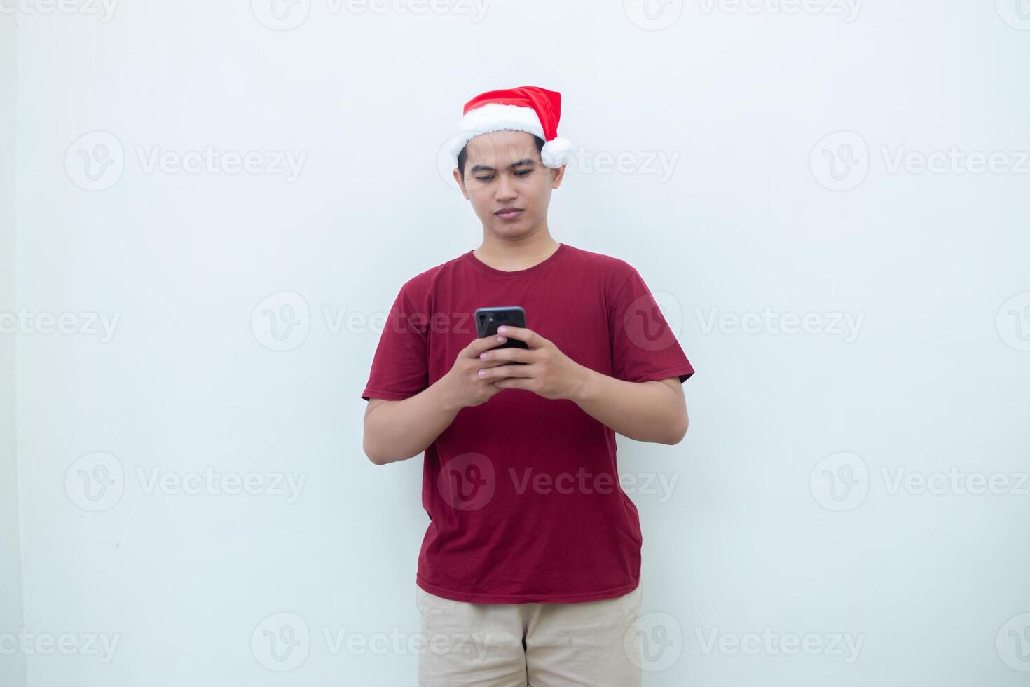 ung asiatisk man bär en santa claus hatt innehav en smartphone och uttrycker en leende, chock och överraskning isolerat förbi en vit bakgrund för visuell kommunikation foto