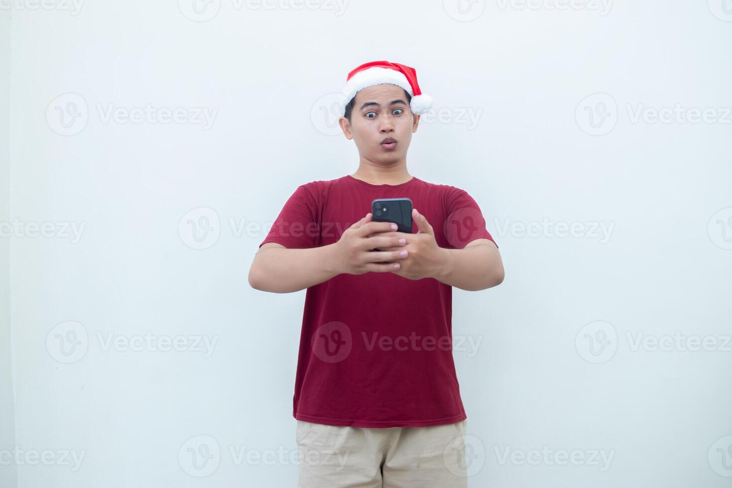 ung asiatisk man bär en santa claus hatt innehav en smartphone och uttrycker en leende, chock och överraskning isolerat förbi en vit bakgrund för visuell kommunikation foto