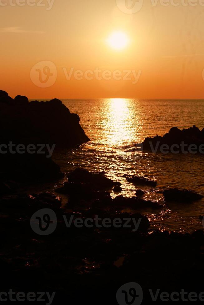 de Sol är miljö på de hav med stenar i de förgrund, svart silhuetter på de stenar, ljus reflekterande av de hav. foto