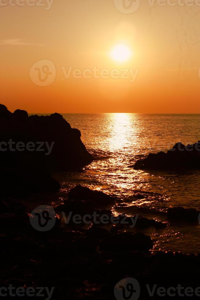 de Sol är miljö på de hav med stenar i de förgrund, svart silhuetter på de stenar, ljus reflekterande av de hav. foto