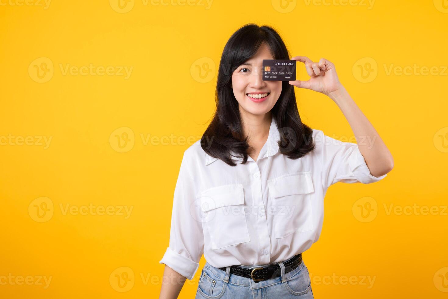 asiatisk ung kvinna innehav kreditera kort i främre av ett öga med Lycklig leende isolerat på gul bakgrund. betalning handla uppkopplad begrepp. foto
