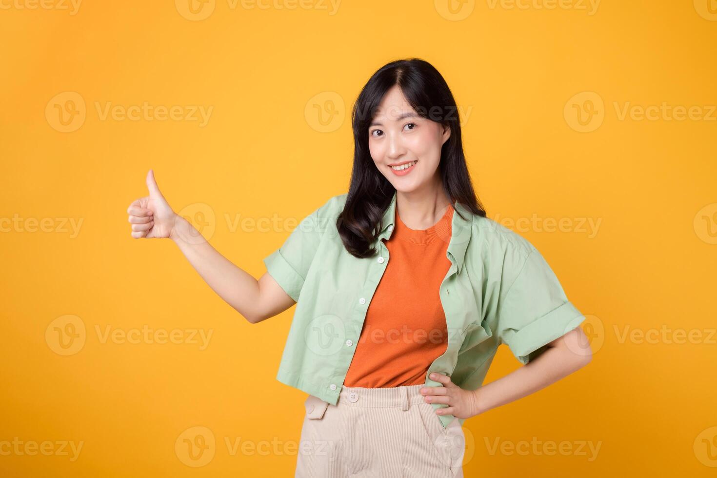 godkännande med ung asiatisk kvinna i henne 30-talet, elegant klädd i ett orange skjorta och grön hoppare. henne tummen upp gest, uppsättning mot en solig gul bakgrund, symboliserar de begrepp av bekräftelse. foto