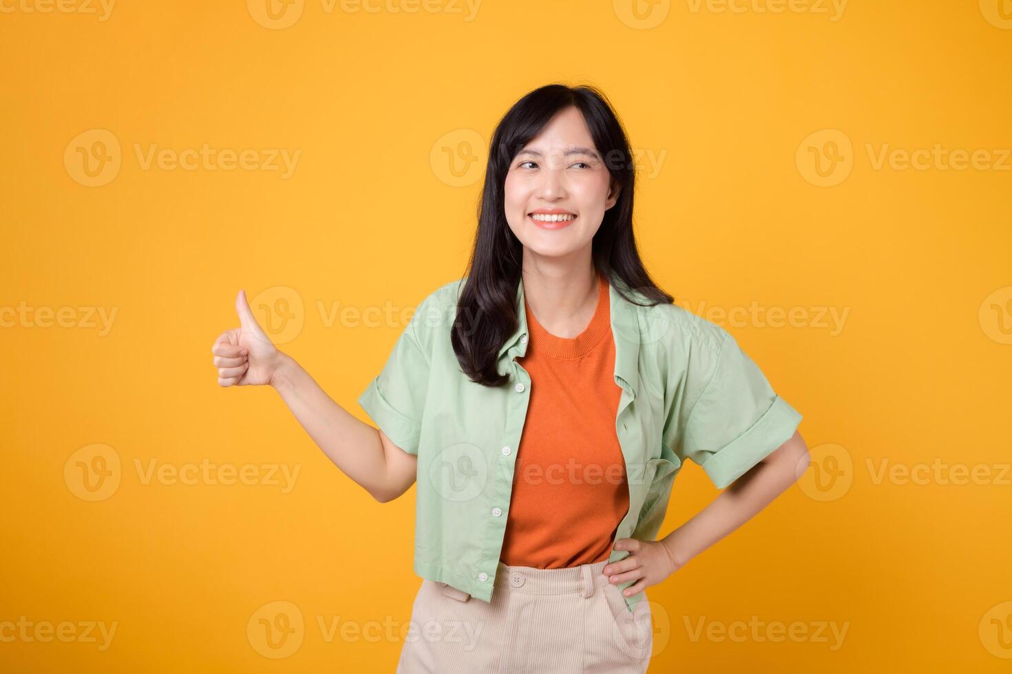 ung asiatisk kvinna i henne 30-talet, påklädning ett orange skjorta och grön hoppare. henne tummen upp gest, isolerat på en vibrerande gul bakgrund, betecknar en begrepp av positivitet och avtal. foto