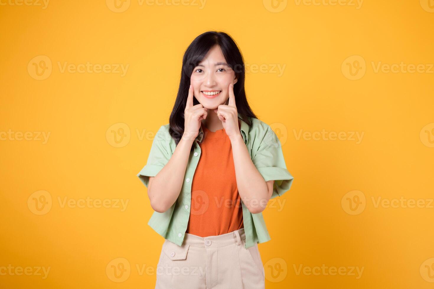 dental vård med ung asiatisk kvinna 30-talet, klädd i orange skjorta och grön hoppare poäng till henne tänder, isolerat på en vibrerande gul bakgrund, highlighting de betydelse av dental sjukvård foto