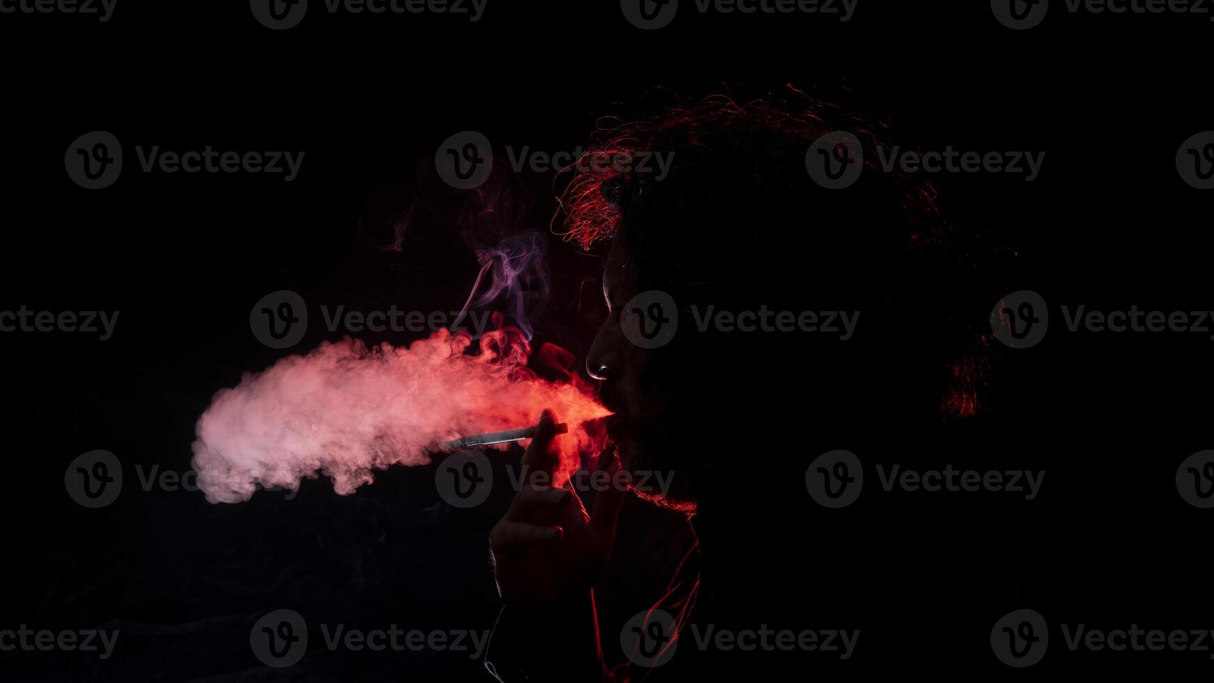 man med en cigarett i hans hand, blåser rök ut av hans mun sett i profil i silhuett med röd ljus belysande hans profil mot svart bakgrund foto