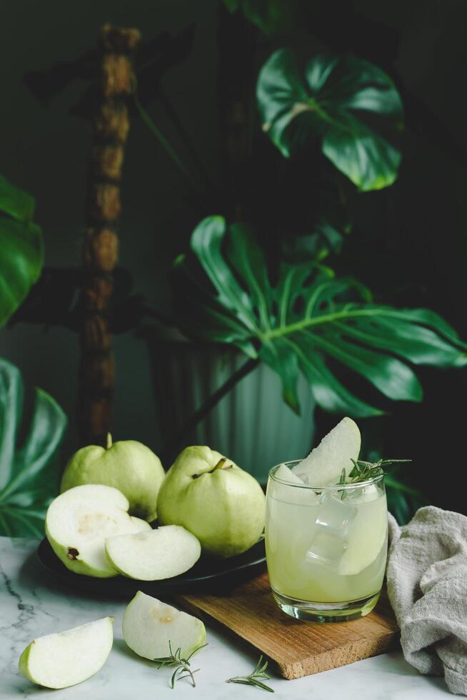 uppfriskande utsökt av iced guava juice i en glas och skivad guava placerad på vit marmor, krukväxt grön växt bakgrund. dekorativ sammansättning drycker på Hem . vertikal Foto