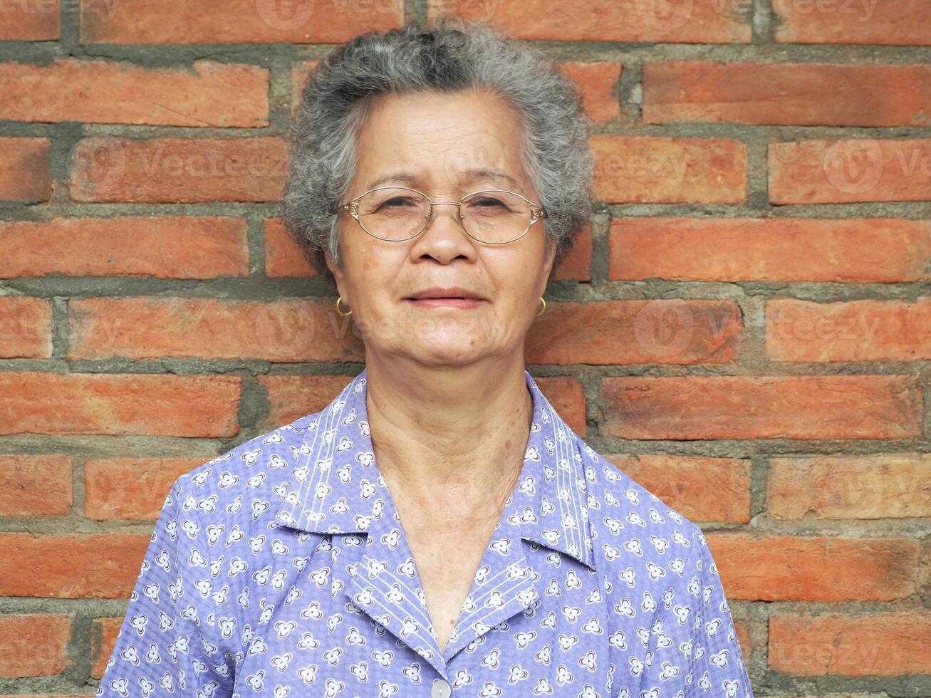 asiatisk äldre kvinna ser på kamera och leende medan stående med tegel vägg bakgrund foto