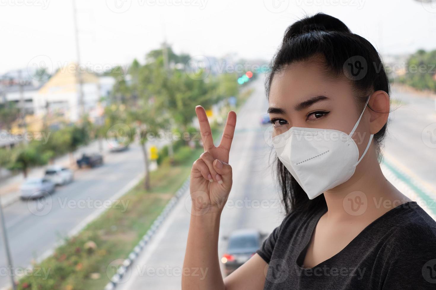 kvinna stående hand tecken på 2 fingrar med att sätta på respirator n95 mask för att skydda mot luftburna luftvägssjukdomar som influensa covid-19 coronavirus pm2.5 damm och smog på vägen burred backgro foto
