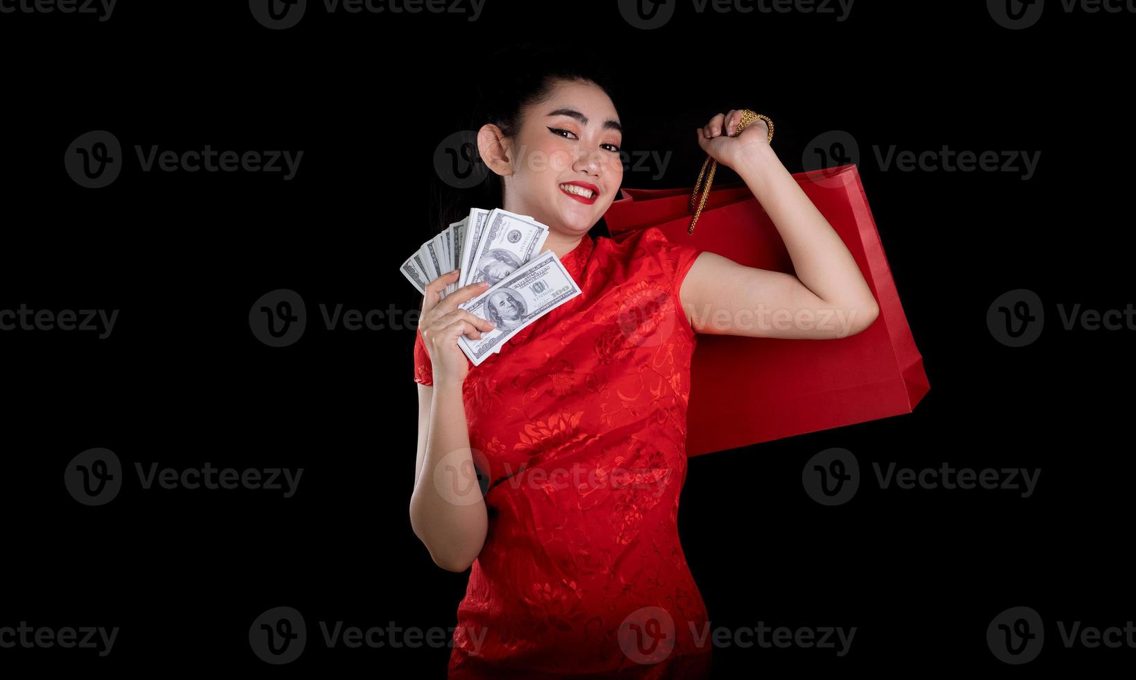 porträtt av en vacker ung asiatisk kvinna röd klänning traditionell cheongsam håller en väska shopping och pengar sedel 100 usd på den svarta bakgrunden foto