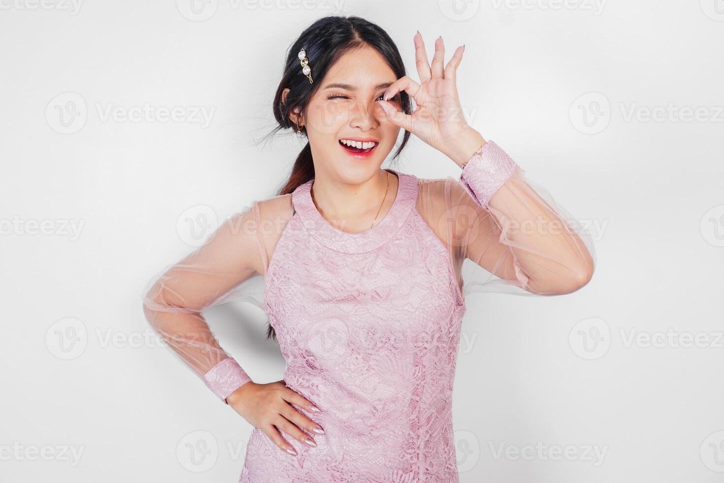 attraktiv ung asiatisk kvinna i rosa klänning är gestikulerar ok tecken med henne fingrar, isolerat förbi vit bakgrund foto