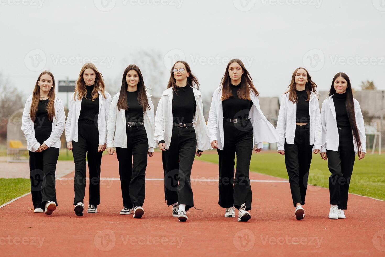 en grupp av många Lycklig Tonårs flickor klädd i de samma utrusta har roligt och Framställ i en stadion nära en högskola. begrepp av vänskap, stunder av lycka. skola vänskap foto