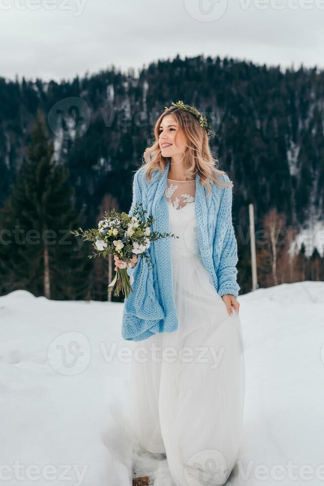 porträtt av en söt brud i en vit klänning med en bukett av blommor i henne händer, insvept i en blå pläd, väntar för de brudgum mot de bakgrund av en vinter- landskap av snötäckt berg. foto