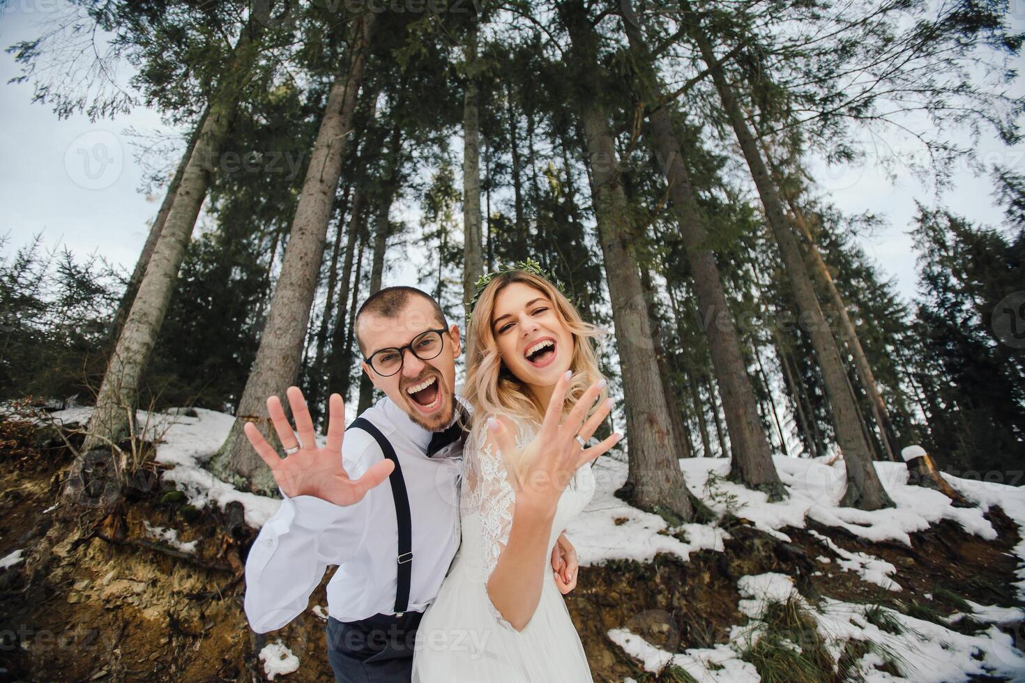 en ung par, de brud och brudgum, leende visa av deras bröllop ringar mot de bakgrund av lång träd. ha en Bra tid skrattande. vinter- bröllop foto