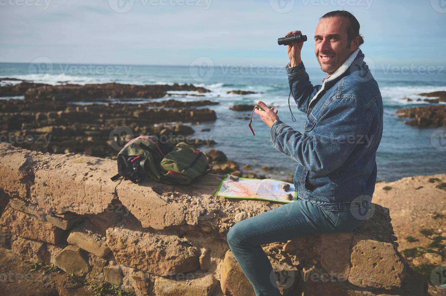 leende turist resande Sammanträde på en sten förbi hav nära hans ryggsäck, innehav kikare, leende ser på de kamera foto