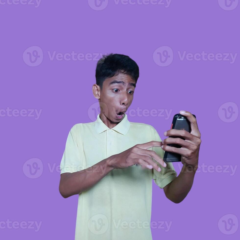 ung asiatisk man överraskad ser på smart telefon, bär gul t-shirt, isolerat lila bakgrund. foto