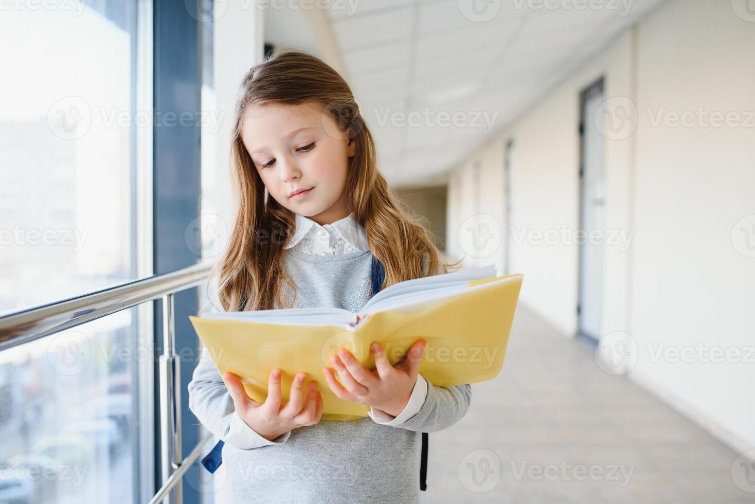 främre se av liten skön skola flicka bland korridor på skola, innehav anteckningar på händer. rolig och Lycklig flicka leende på kamera, vilar efter lektioner på primär skola foto