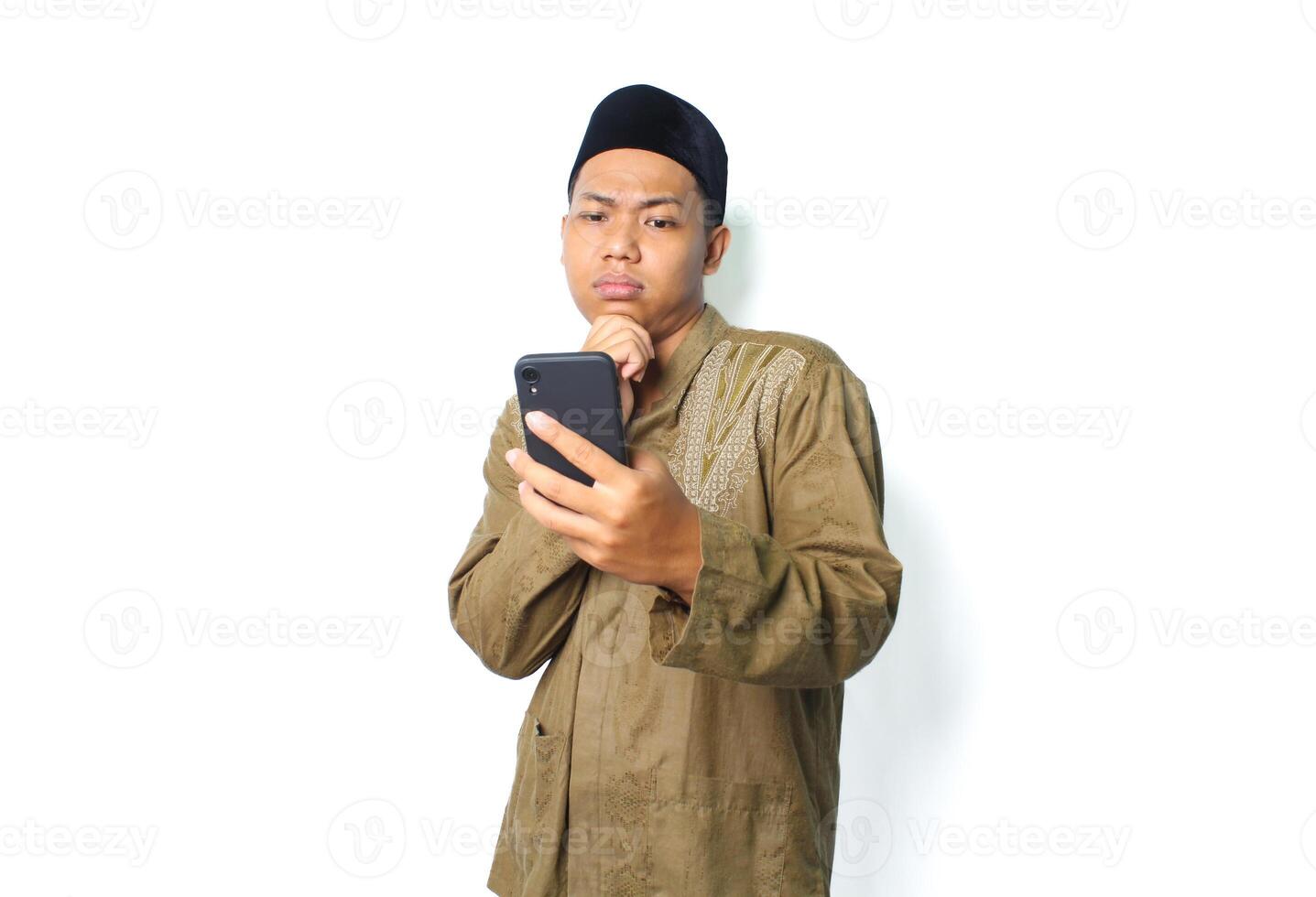 omtänksam asiatisk muslim man innehav mobil telefon med funderar hans haka isolerat på vit bakgrund foto
