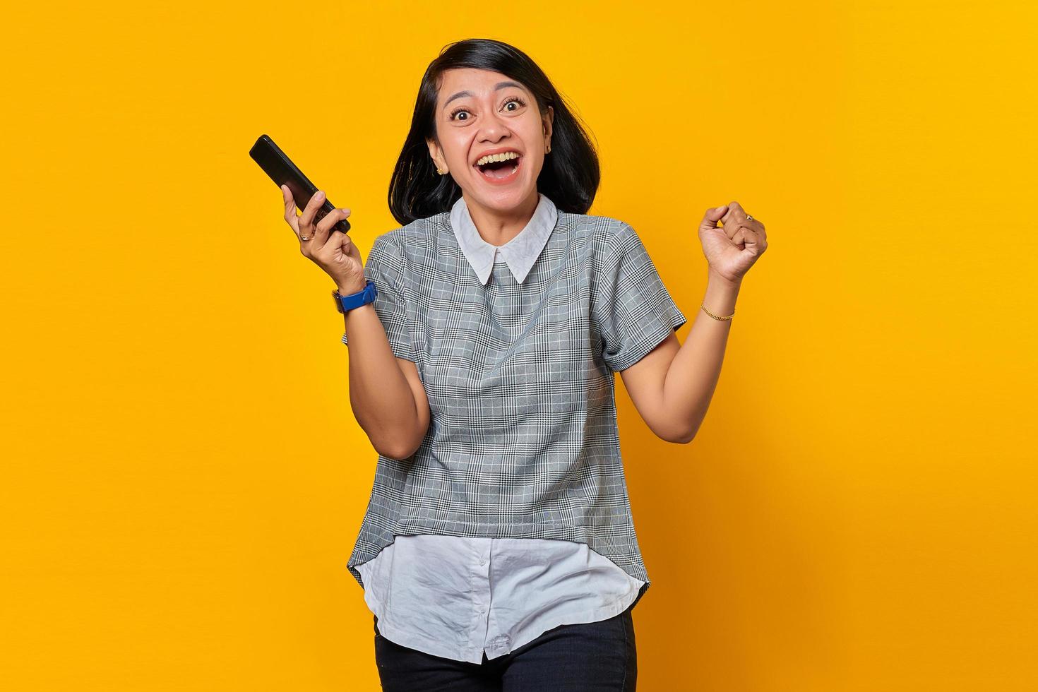 ung asiatisk kvinna känner sig upprymd och glad när hon tittar på meddelanden på smartphone på gul bakgrund foto