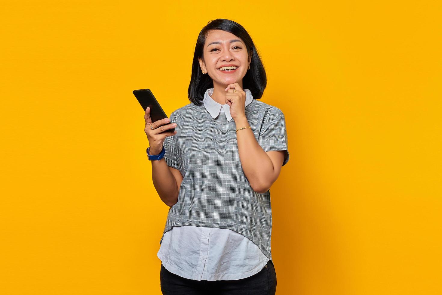 glada leende ung asiatisk kvinna med finger på hakan och hålla mobiltelefon över gul bakgrund foto