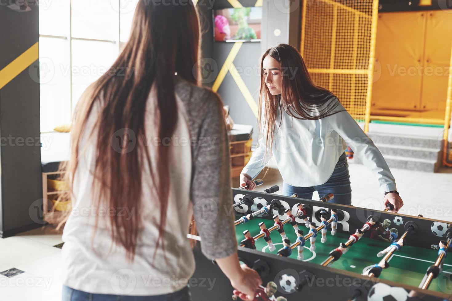 två vackra tvillingflickor spelar bordsfotboll och har kul. en av systrarna håller en leksaksboll i handen och visar tungan foto