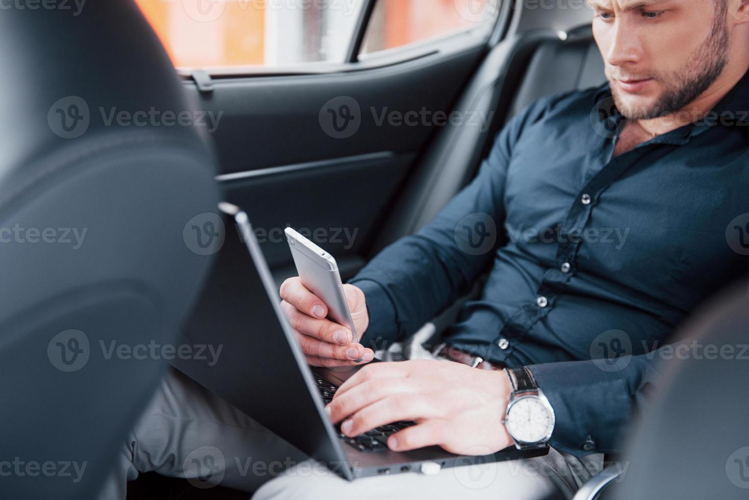 en ung affärsman som arbetar på bärbar dator och pratar i telefon medan han sitter i bilens rygg. arbetar i rörelse, uppskattar sin tid foto