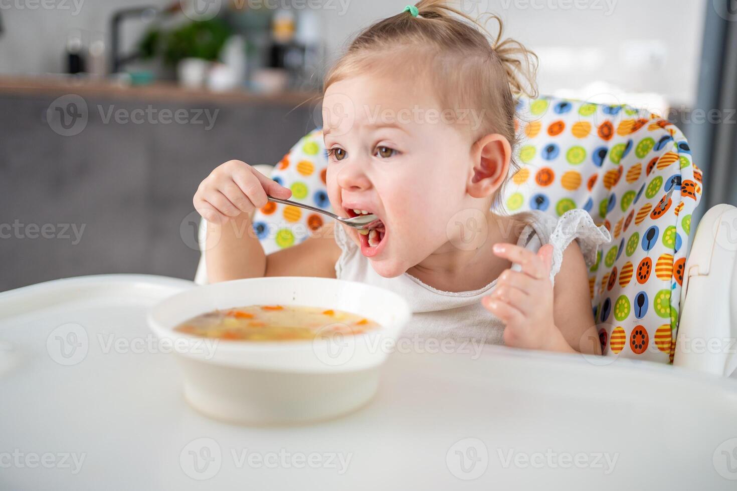 söt bebis flicka litet barn Sammanträde i de hög stol och äter henne lunch soppa på Hem kök. foto