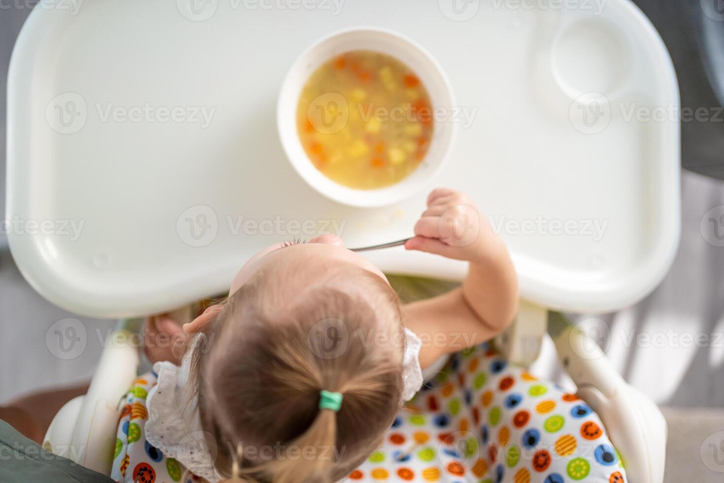 söt bebis flicka litet barn Sammanträde i de hög stol och äter henne lunch soppa på Hem kök. foto