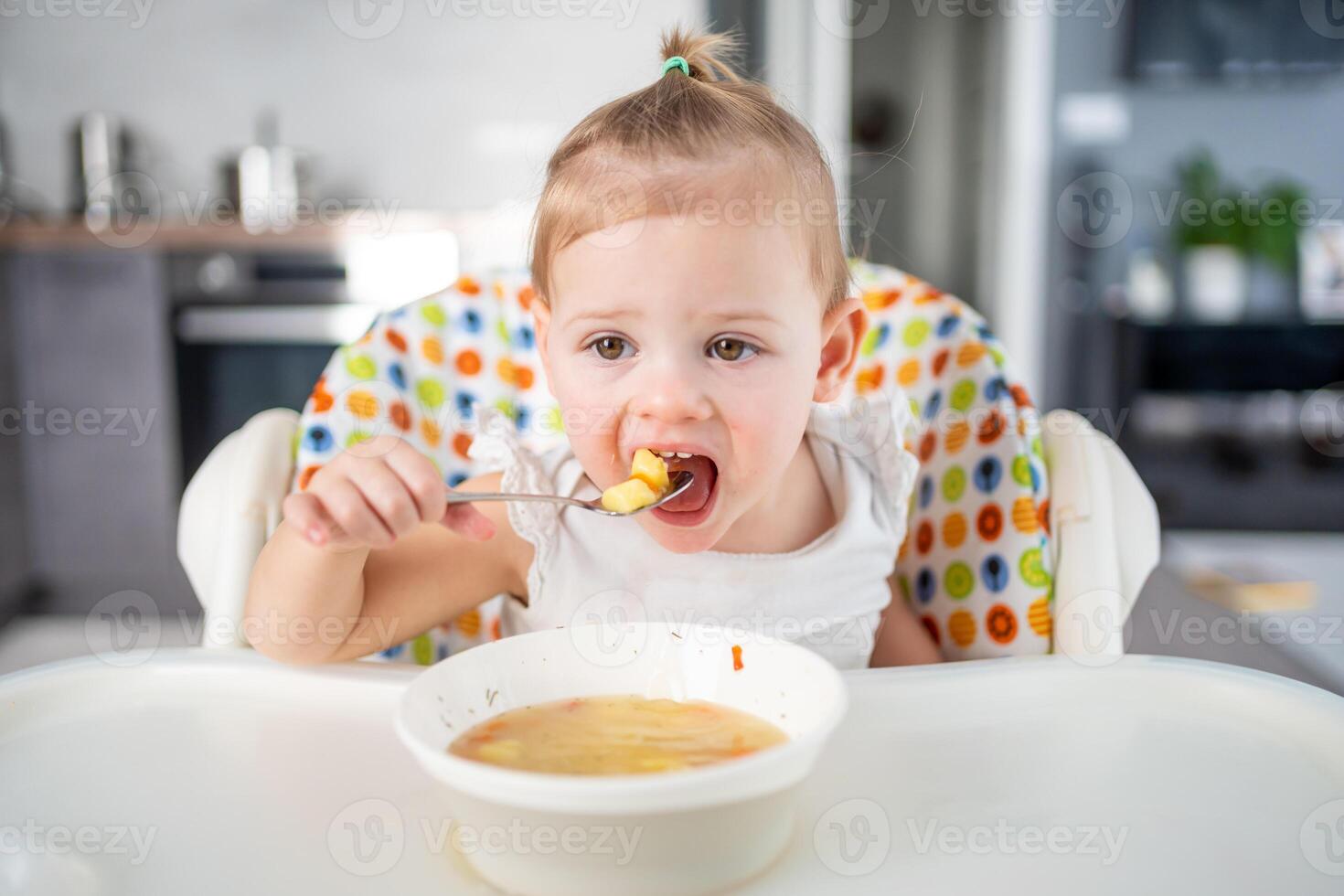 söt bebis flicka litet barn Sammanträde i de hög stol och äter henne lunch soppa på Hem kök. foto