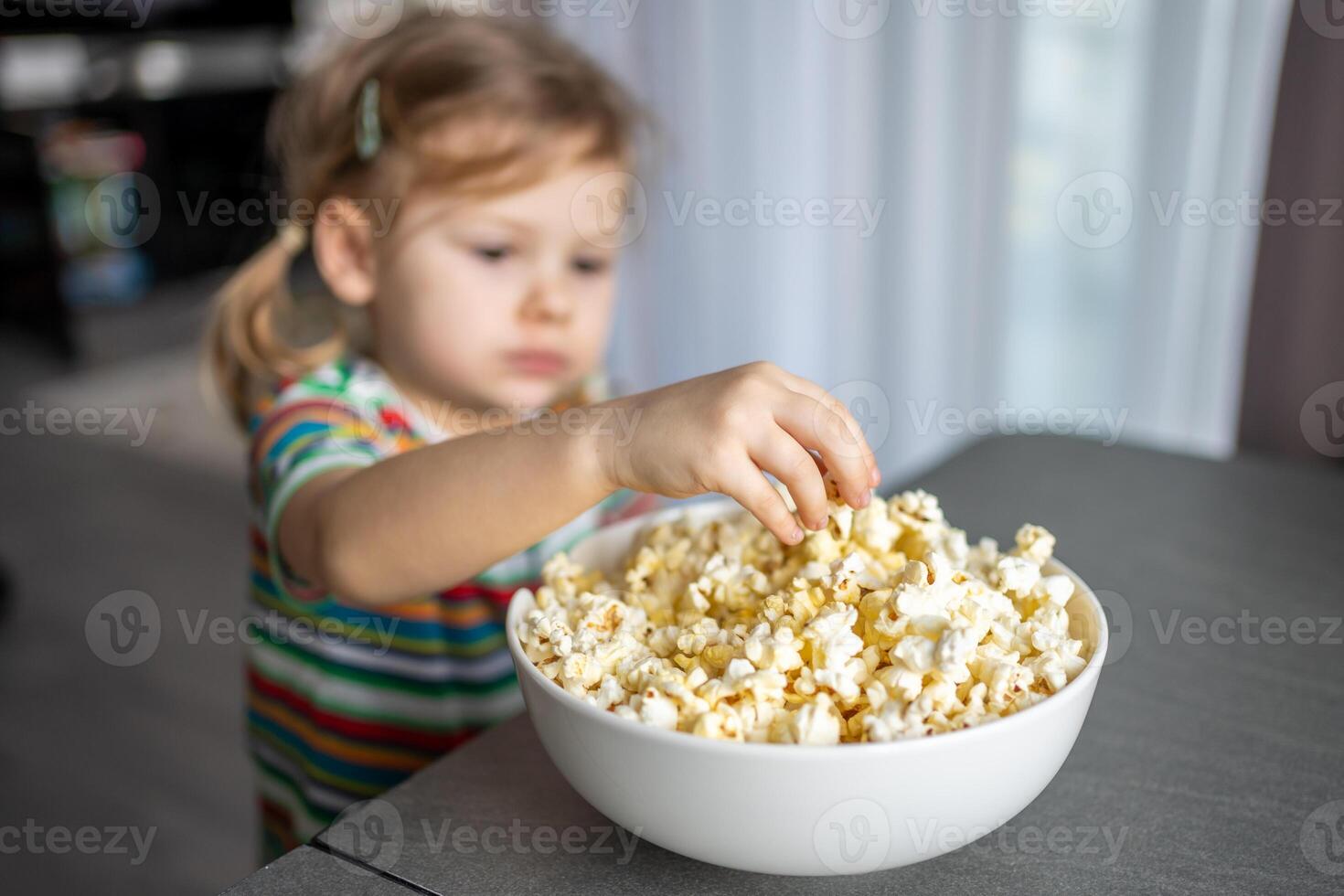 liten flicka är äter popcorn i Hem kök. fokus på hand tar popcorn foto