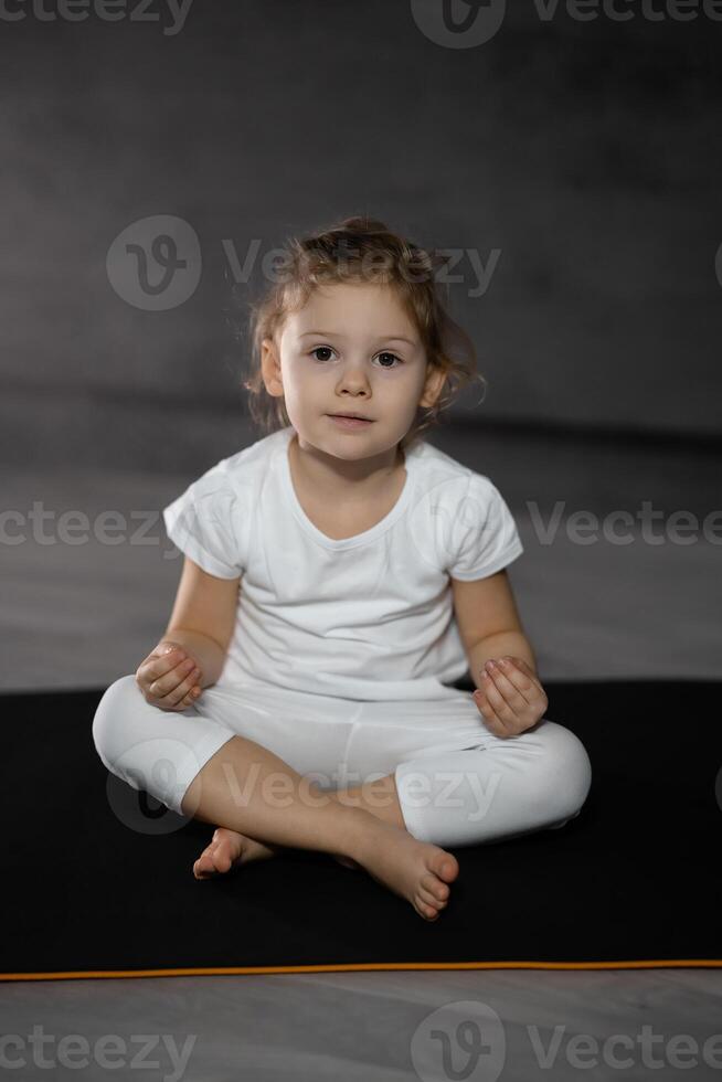 tre år gammal liten flicka mediterar i en lotus utgör på en grå bakgrund i mörk rum. hög kvalitet Foto
