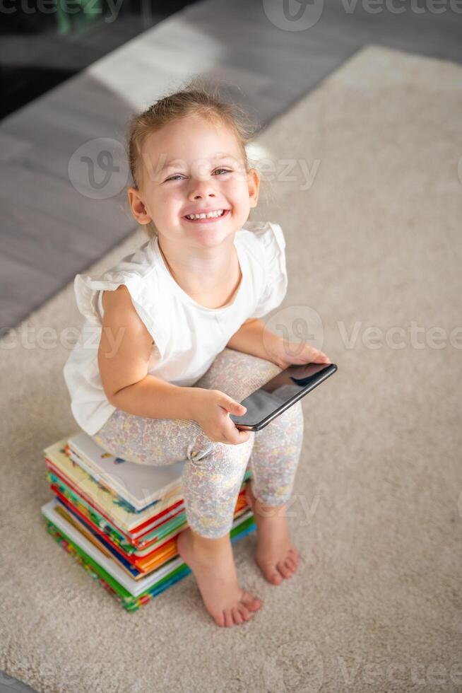 leende liten flicka sitter på stack av barns saga böcker med smartphone i henne händer foto