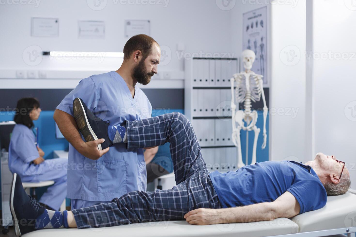 ortopedisk sjuksköterska assisterar senior patient återhämtar sig från knä skada på klinik. tryck lindra osteopatisk behandling förbi kiropraktor för pensionerad man upplever muskulös ömhet i ben. foto