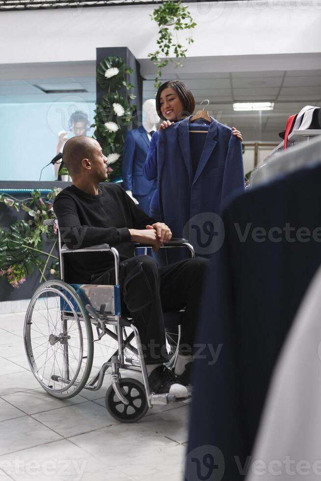 Kläder Lagra asiatisk kvinna anställd tillhandahålla Stöd till kund med fysisk nedsättning i väljer jacka. köpcenter arbetstagare hört arab man i rullstol i välja formell utrusta foto