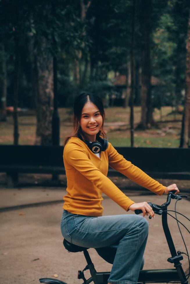 Lycklig ung asiatisk kvinna medan ridning en cykel i en stad parkera. hon log använder sig av de cykel av transport. miljömässigt vänlig begrepp. foto