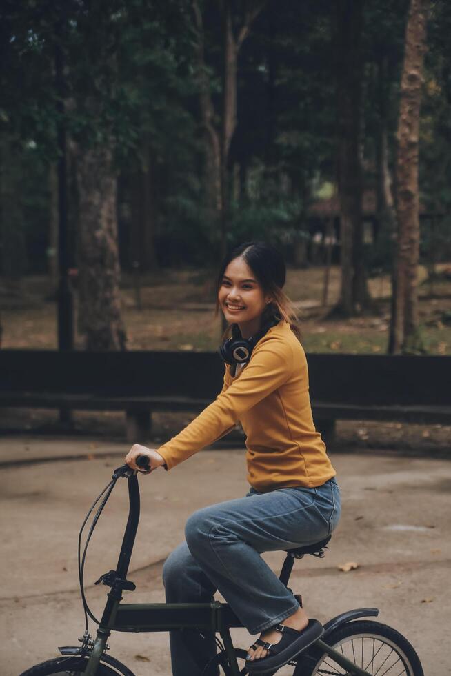 Lycklig ung asiatisk kvinna medan ridning en cykel i en stad parkera. hon log använder sig av de cykel av transport. miljömässigt vänlig begrepp. foto