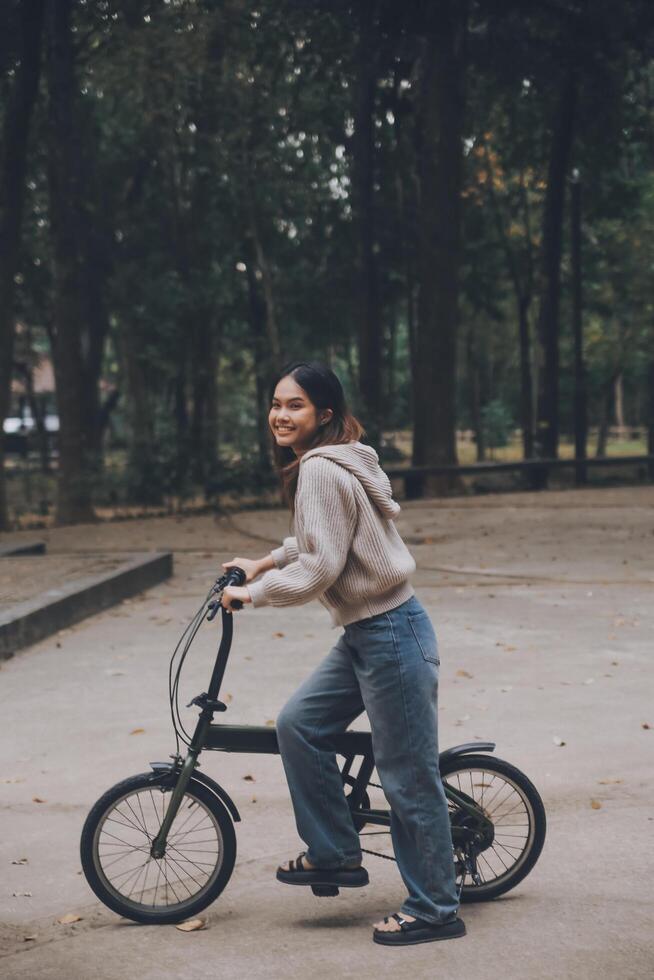 Lycklig ung asiatisk kvinna medan ridning en cykel i en stad parkera. hon log använder sig av de cykel av transport. miljömässigt vänlig begrepp. foto