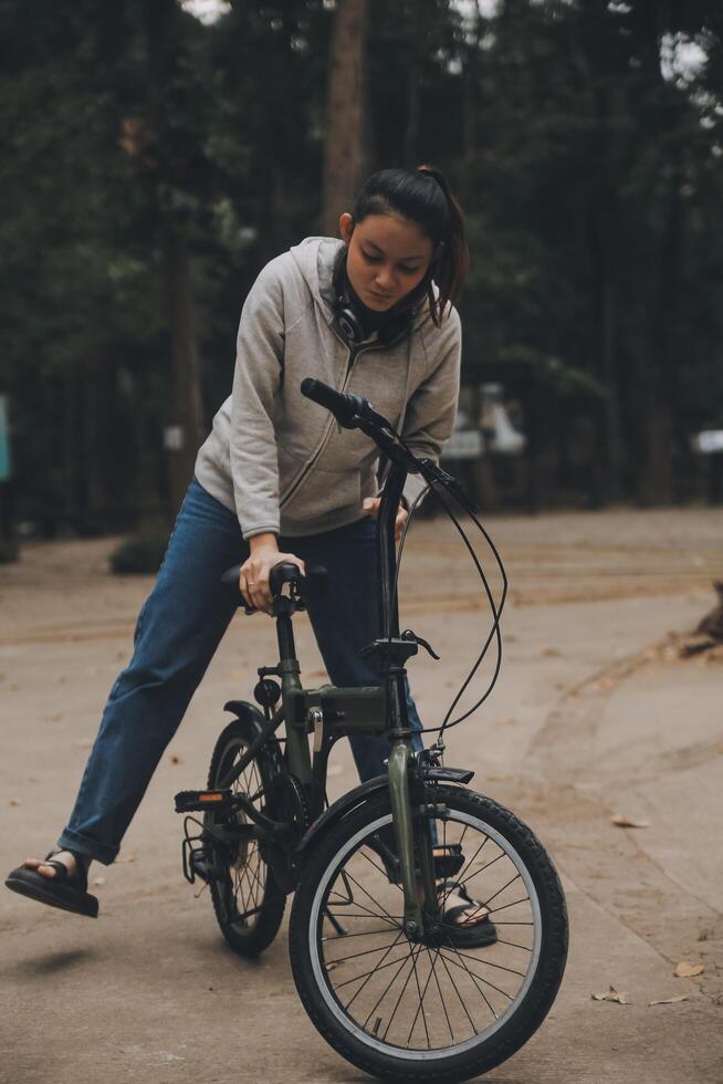 Lycklig asiatisk ung kvinna promenad och rida cykel i parkera, gata stad henne leende använder sig av cykel av transport, eco vänlig, människor livsstil begrepp. foto