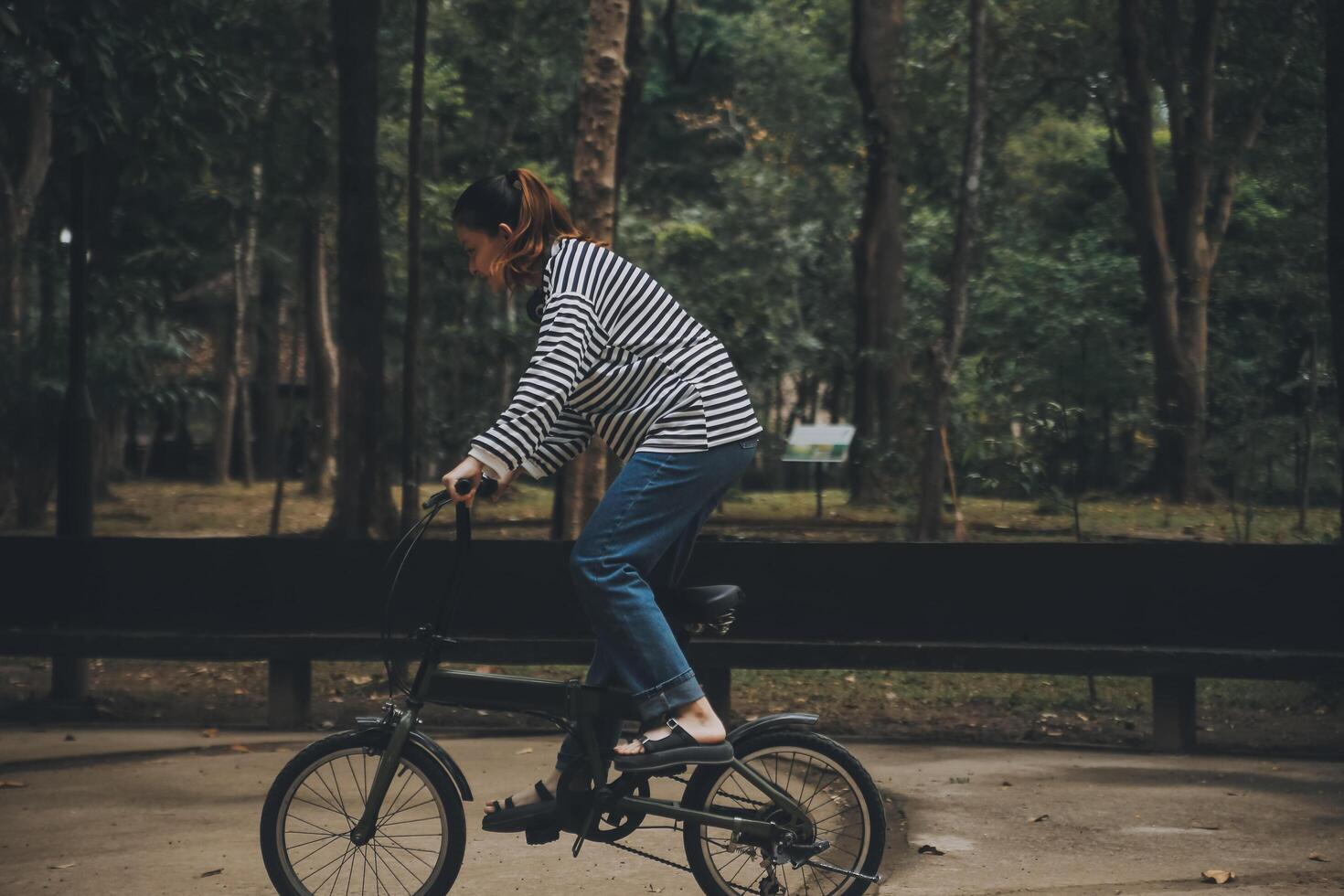 Lycklig asiatisk ung kvinna promenad och rida cykel i parkera, gata stad henne leende använder sig av cykel av transport, eco vänlig, människor livsstil begrepp. foto