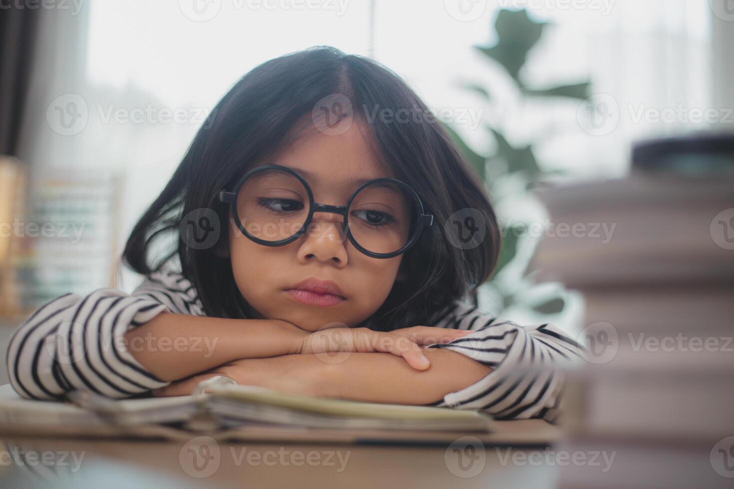 liten asiatisk tjej som sitter ensam och tittar ut med ett uttråkat ansikte, förskolebarn som lägger huvudet på bordet med ledsen uttråkad med läxor, bortskämt barn foto