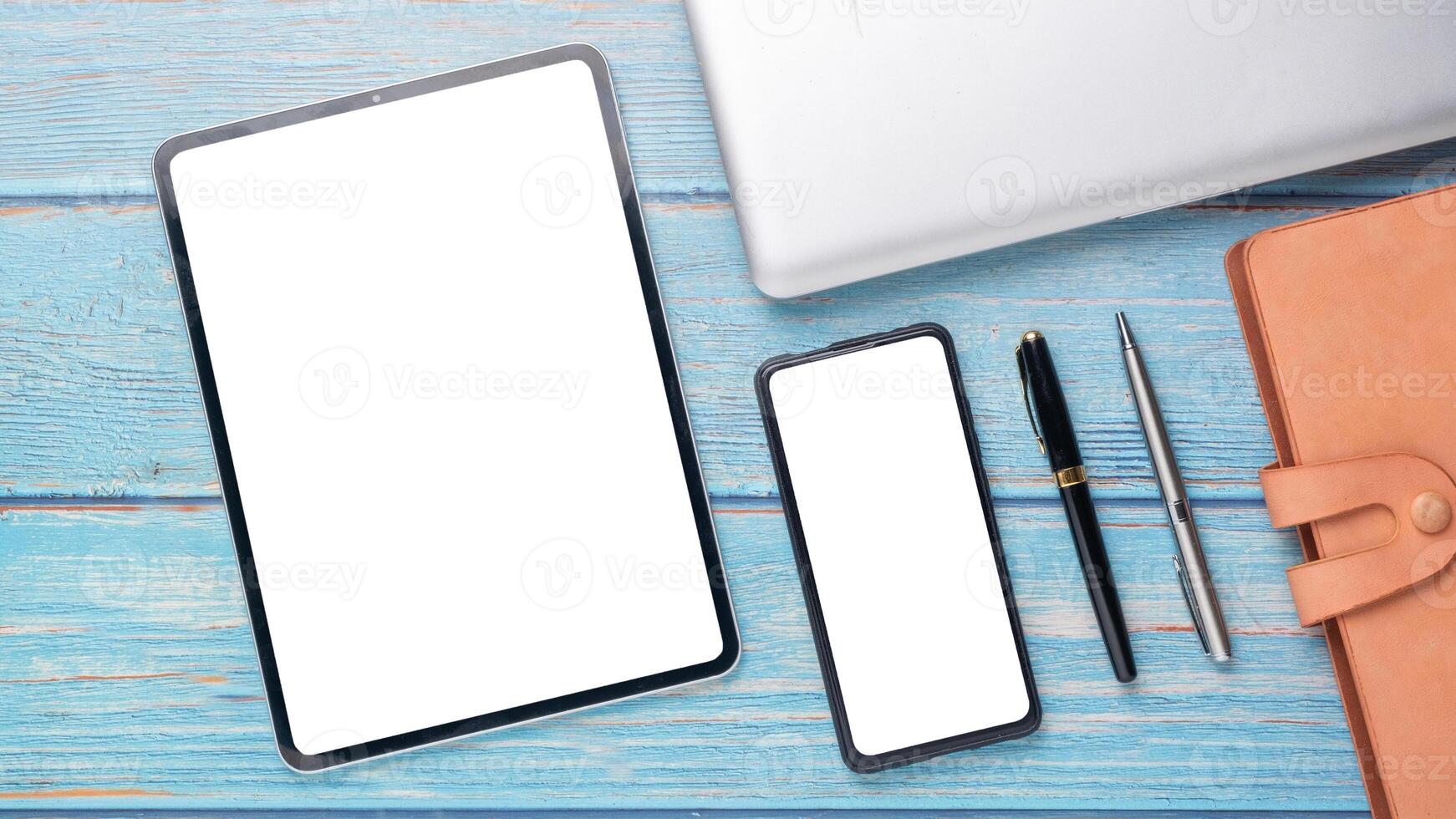modern professionell arbetsyta terar en läsplatta och smartphone med tom skärmar, vid sidan av elegant pennor och en läder anteckningsbok på en blå trä- yta. foto