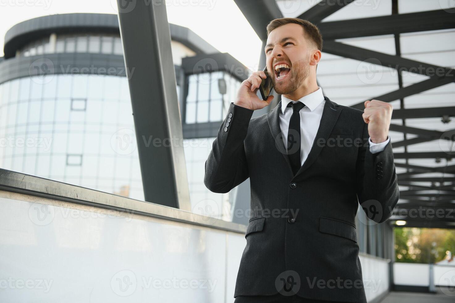 affärsman leende och höjning hans näve i de luft, med kontor byggnad bakgrund - företag Framgång, prestation, och vinna begrepp foto
