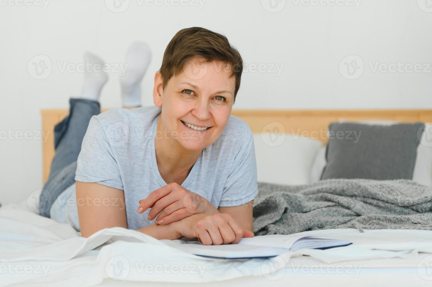 mitten ålder blond kvinna läsning bok liggande på de säng på Hem. foto