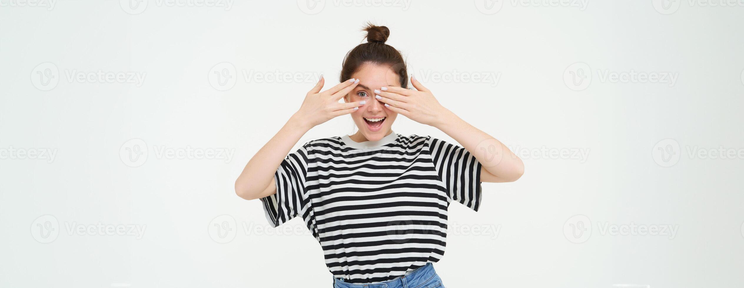 Lycklig ung kvinna, födelsedag flicka kikar genom fingrar, innehar händer på ögon, väntar för överraskning, står över vit bakgrund foto