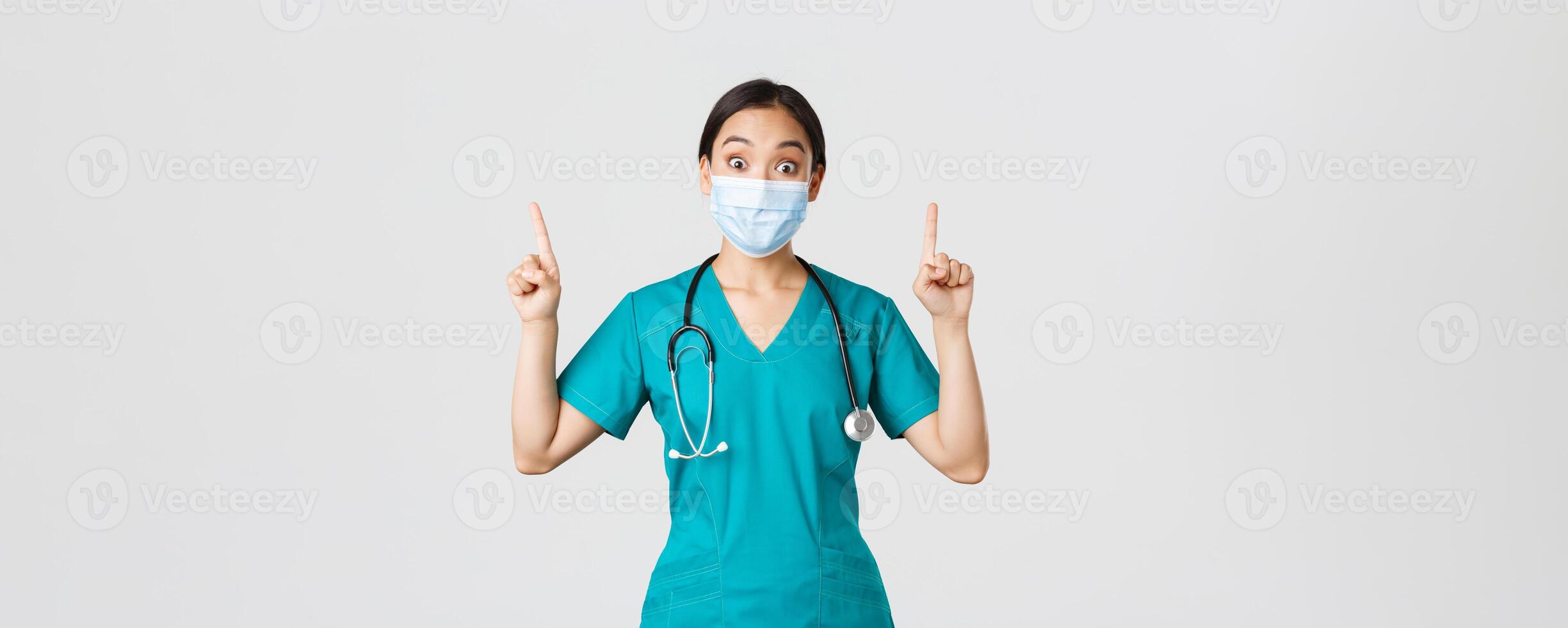 covid19, coronavirus sjukdom, sjukvård arbetare begrepp. fascinerad asiatisk kvinna läkare, läkare eller internera i medicinsk mask och skrubbar, ser nyfiken, pekande fingrar upp, vit bakgrund foto